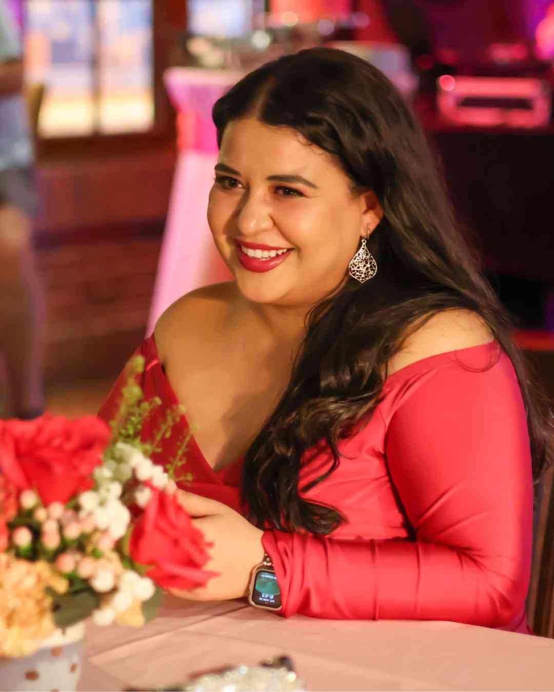 A smiling woman in a red off-shoulder dress and dangling earrings sits at a table with red and white flowers at an evening party.