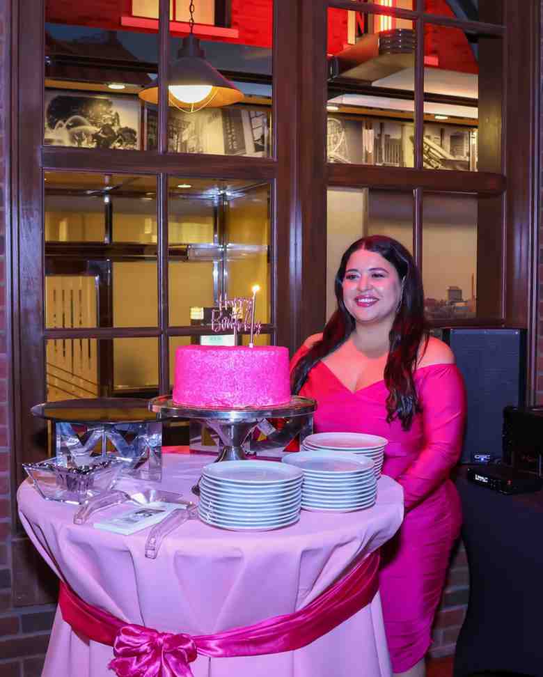 A woman in a bright pink dress stands beside a table with a sparkly pink birthday cake and stacked plates, decorated with a pink bow.