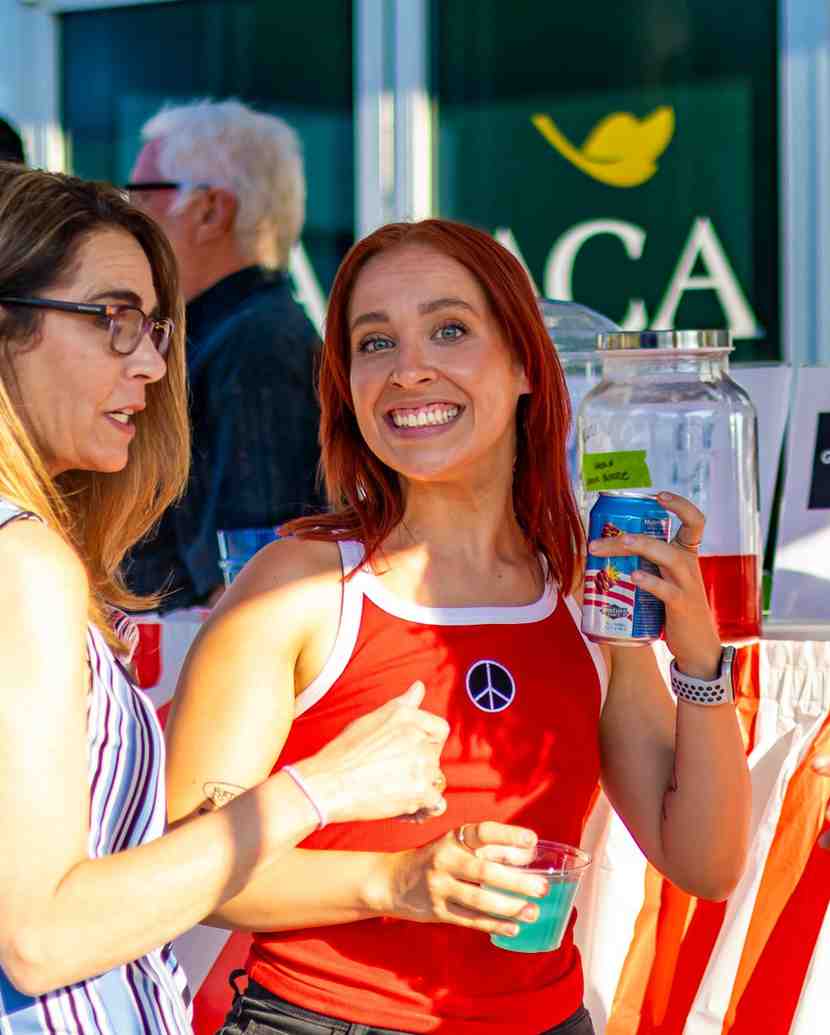 A smiling woman in a red tank top holds a drink can and cup while talking with another woman at an outdoor event with a banner behind them.