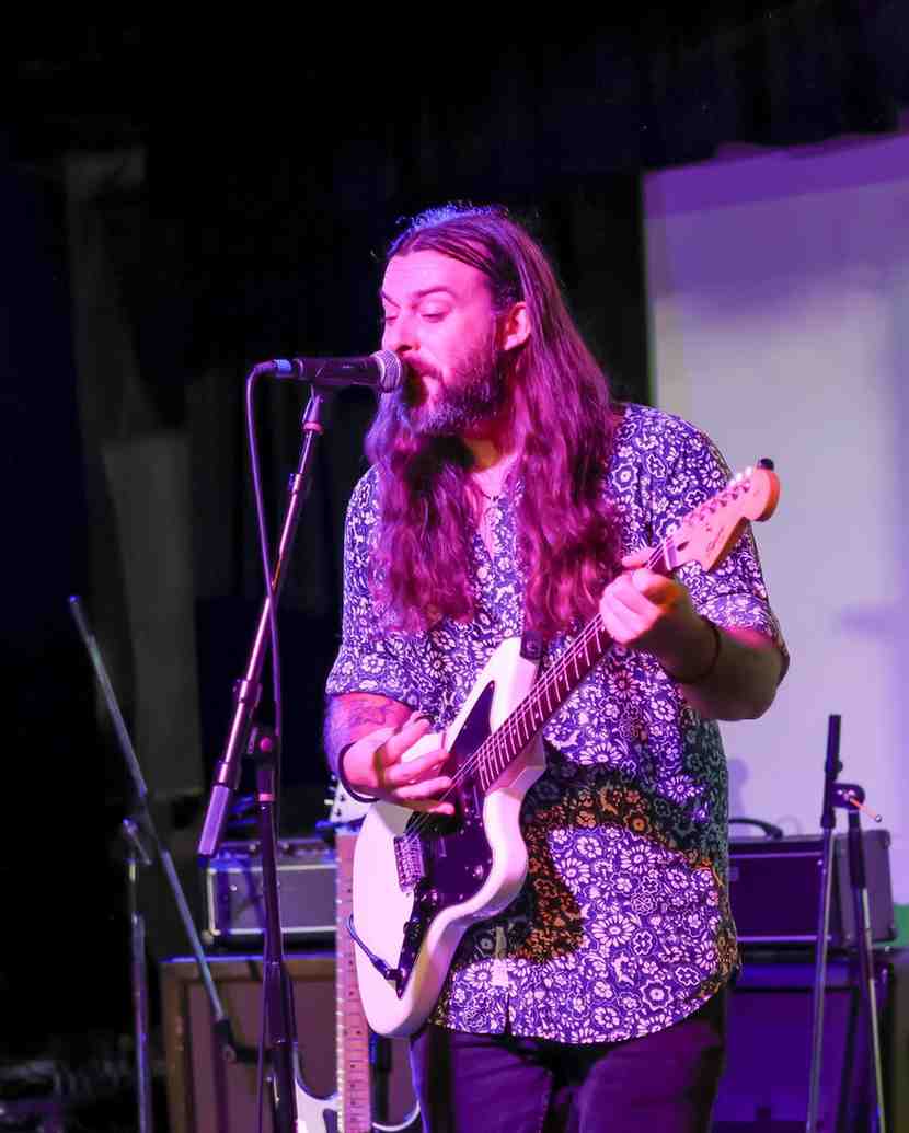 A long-haired man sings into a microphone while playing an electric guitar on stage, lit by purple stage lighting.