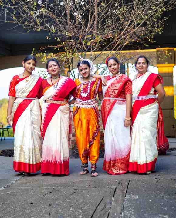 Five women in traditional red and white Indian sarees and one in an orange dance costume pose together outdoors under bare tree branches.