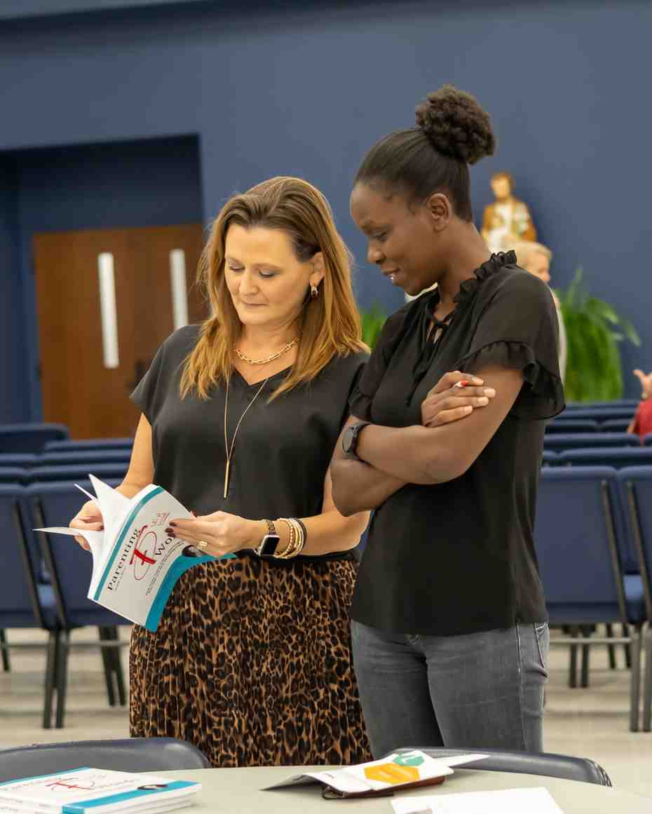Two women look together at an open copy of the book Parenting with a Few F Words in a room with rows of blue chairs.
