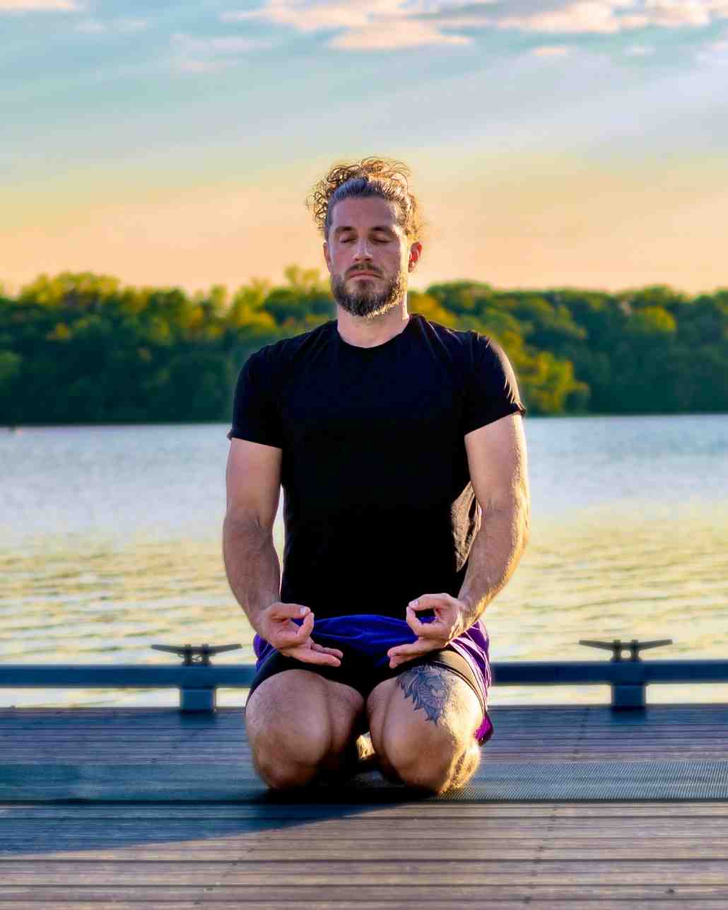 A bearded man kneels in a meditation pose with eyes closed on a wooden dock beside a lake at sunset.
