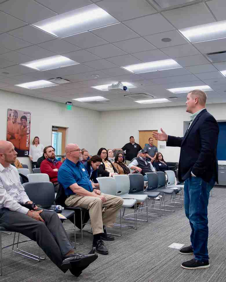 A man in a blazer speaks and gestures to a seated group in a room with a 'Small Business' poster on the wall.