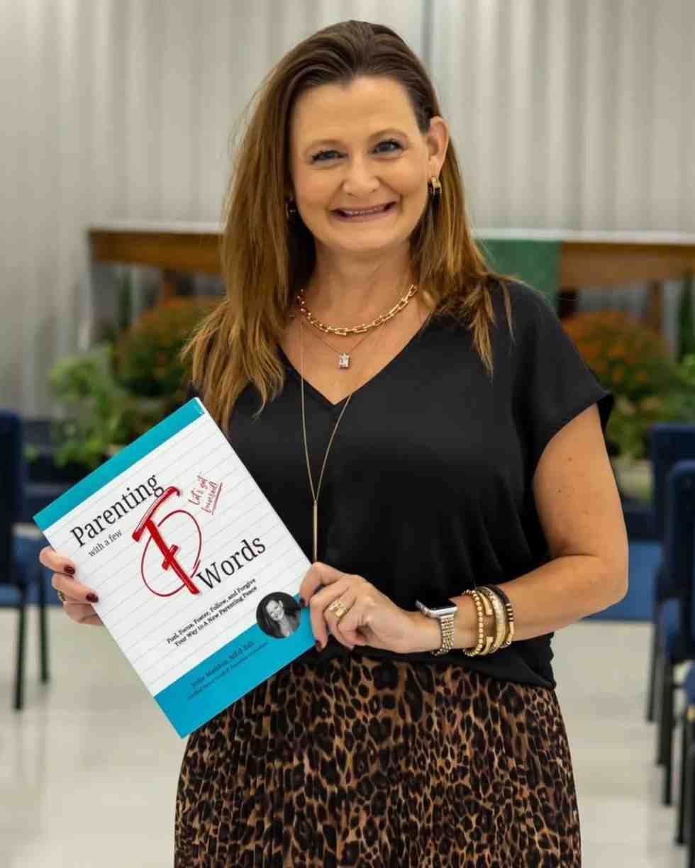A woman in a black top and leopard-print skirt smiles while holding up a paperback book titled Parenting with a Few F Words.