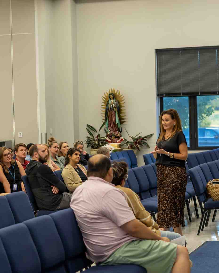 A woman in a black top and leopard-print skirt speaks and gestures to a seated audience in a room with a religious statue in the corner.