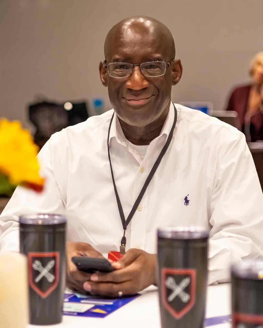 A smiling man in glasses and a lanyard sits at a conference table with a phone, flanked by tumblers printed with a crossed-wrench shield logo.