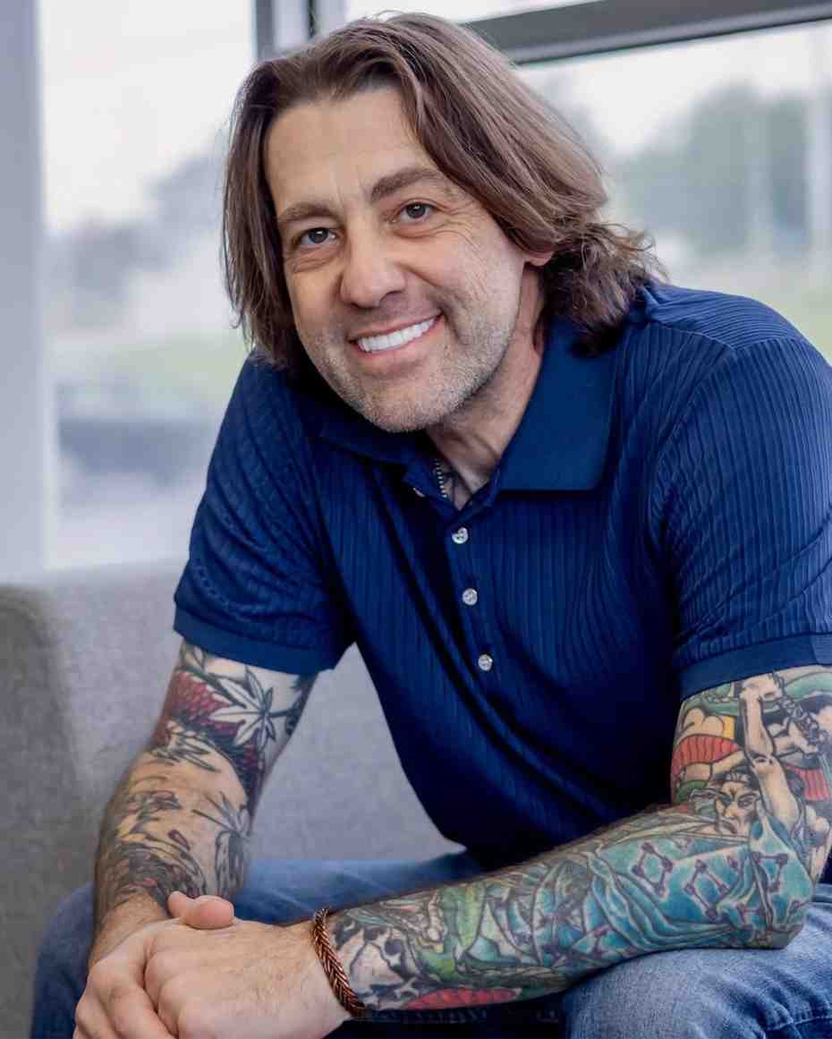 A smiling man with long hair, stubble, and full sleeve tattoos leans forward with clasped hands, seated by a bright window.