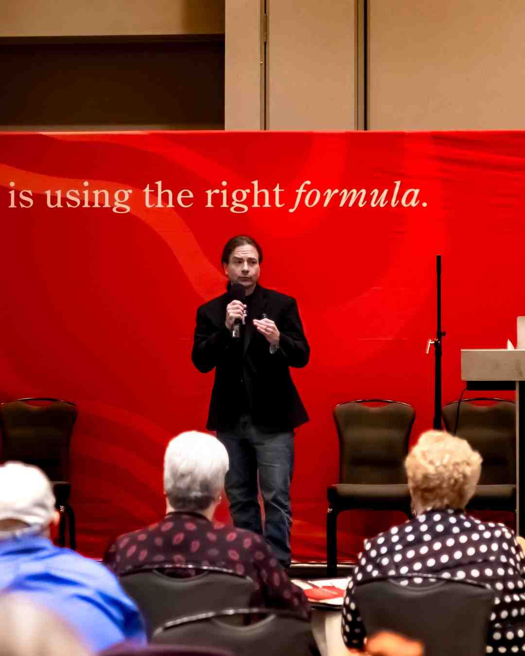 A man holds a microphone and speaks on stage in front of a large red banner reading 'is using the right formula', with seated attendees in the foreground.