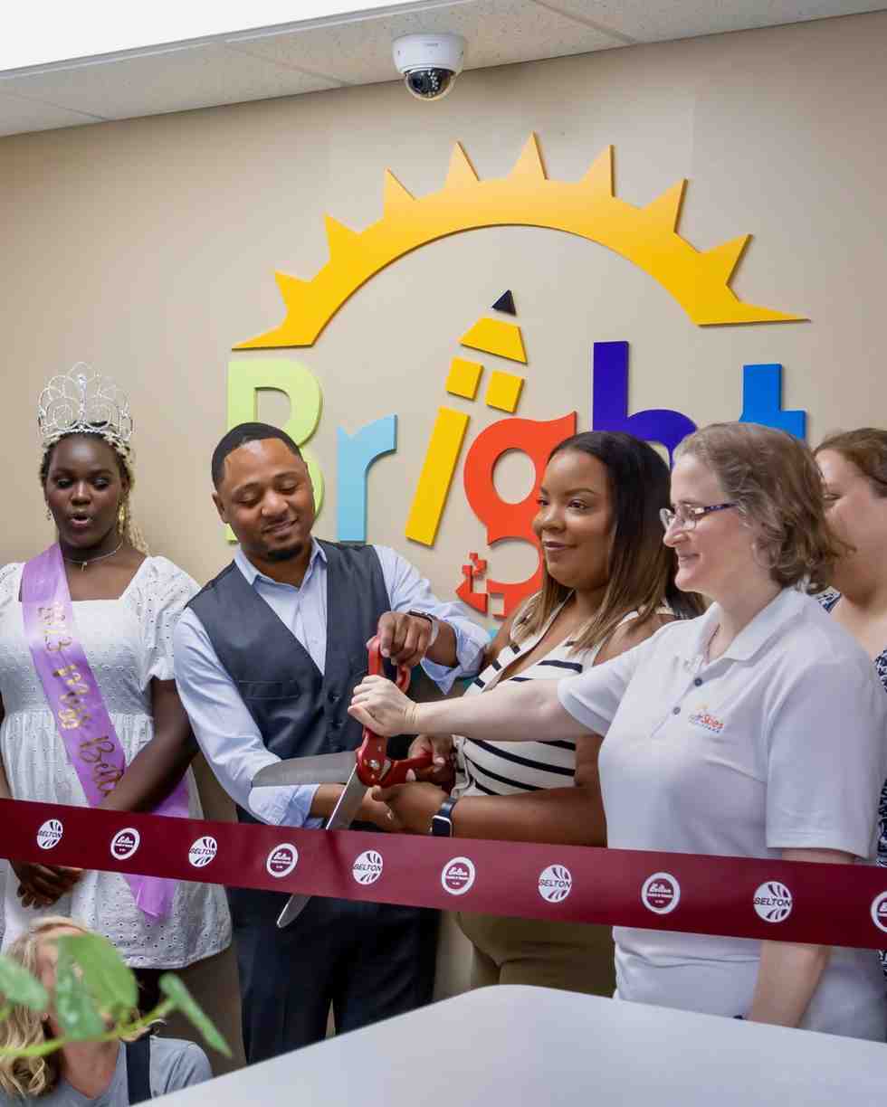 A group of people cut a ribbon together in front of a colorful sunrise 'Bright' logo mural, one woman wearing a pageant sash and tiara.